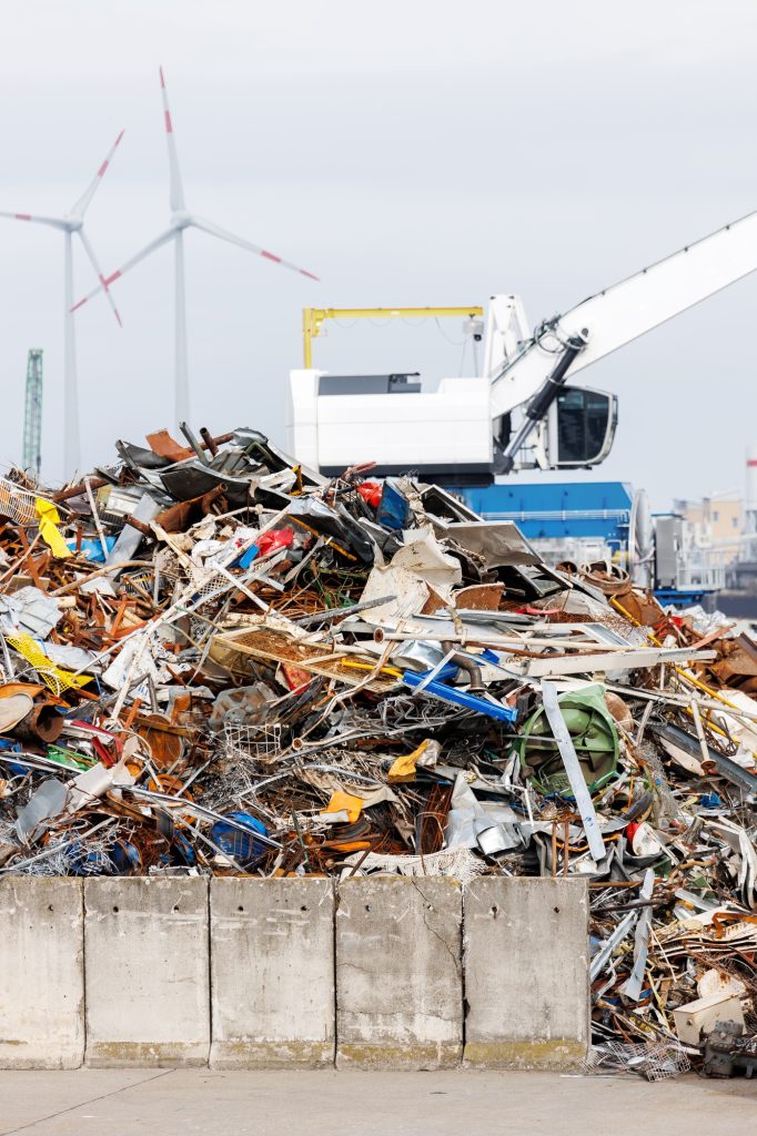 Crane at recycling facility loading large scrap metal pile with wind turbines in background.